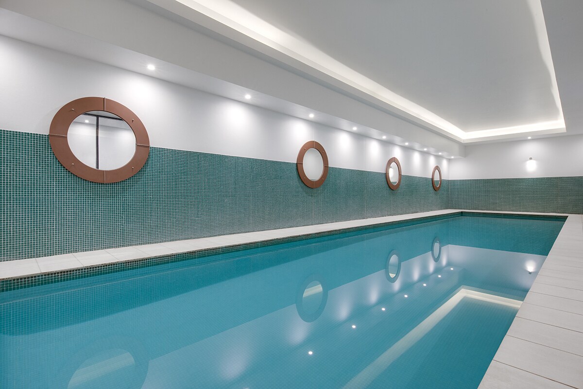 An indoor pool area is presented with a smooth, reflective surface. The walls are adorned with circular mirrors and feature a teal tiled accent. Soft lighting highlights the space, creating a serene swimming environment.