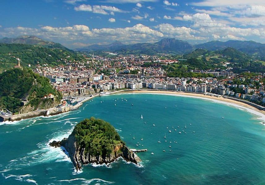A panoramic view captures a vibrant coastal town nestled along a crescent-shaped bay. Gentle waves lap against the sandy shore, while boats are seen dotting the water. Lush green hills frame the town, and clouds drift across a bright blue sky.