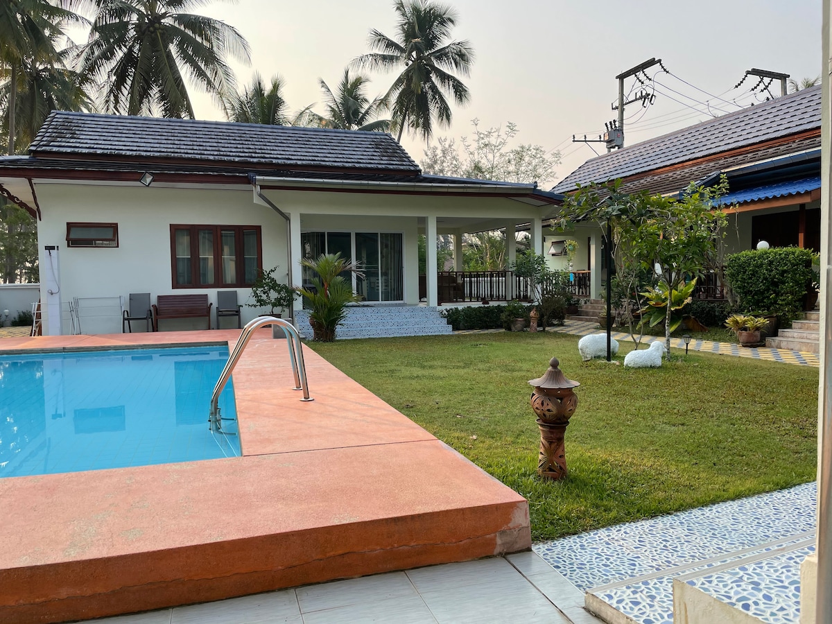 The outdoor area features a swimming pool surrounded by manicured grass and palm trees, offering a tranquil environment. A large terrace with seating is visible, along with a well-maintained bungalow exterior that includes sliding doors and a welcoming entryway.