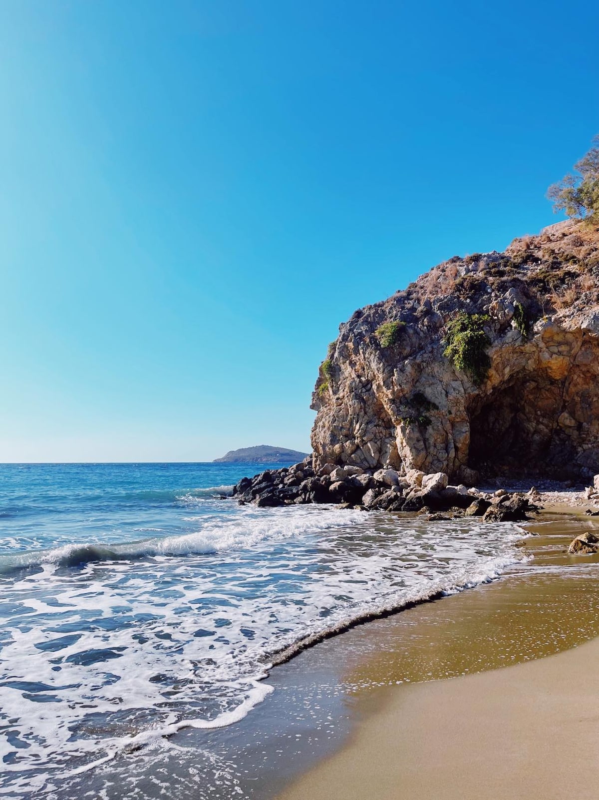A sandy beach is framed by rocky cliffs under a clear blue sky. Gentle waves lap against the shore, creating a serene atmosphere. The landscape showcases natural elements, with vegetation creeping along the rocky formations and distant hills visible along the horizon.