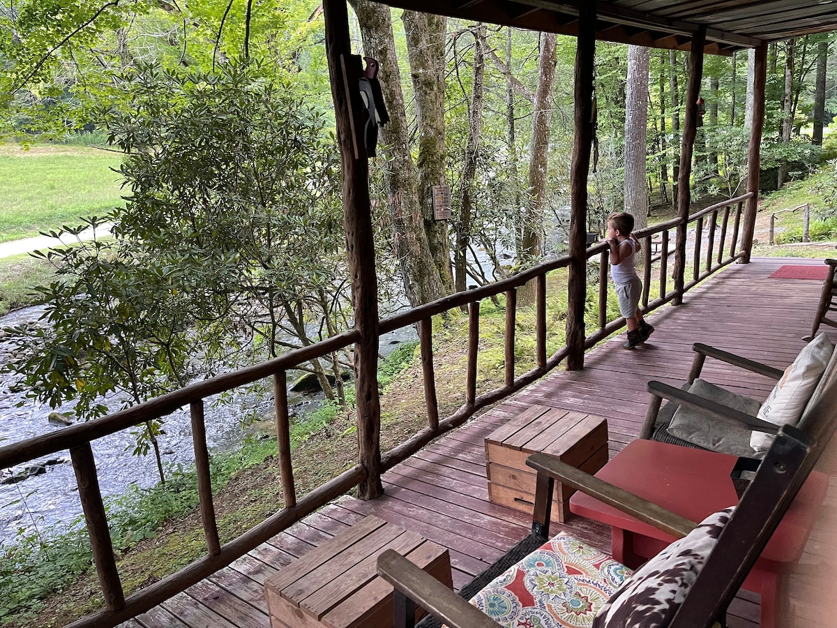 A covered porch features rustic wooden railing and seating options, providing a view of the creek and surrounding trees. The wooden floor is complemented by an area rug, while a child can be seen observing the scenery, highlighting the connection with nature.