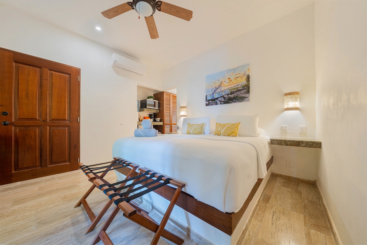 A contemporary guest room features a spacious king size bed with neutral bedding and decorative pillows. A wooden ceiling fan hangs above, while a wall-mounted artwork adds a pop of color. A kitchenette is visible in the background, and light streams through the door leading outside.