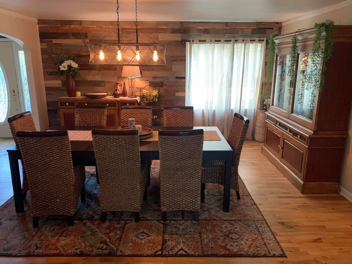 A dining area is presented with a large wooden table surrounded by six woven chairs. A rustic accent wall with varied wood tones creates warmth. Soft lighting is provided by a multi-bulb fixture, while a sideboard and decorative elements add depth to the space.