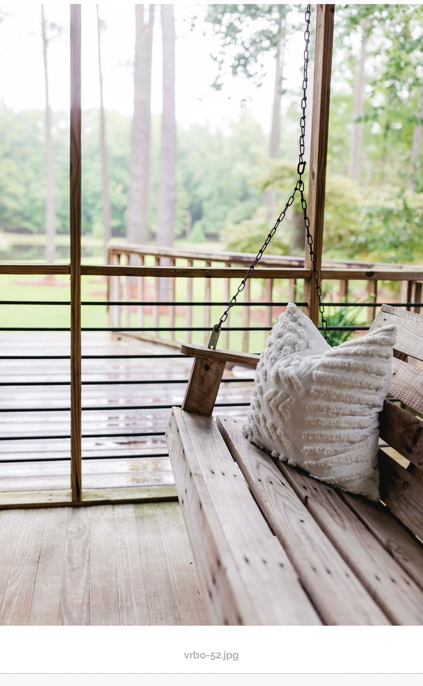 A wooden swing sits on a covered porch, offering a comfortable spot to relax. A textured white pillow adds a soft touch. Lush greenery and tall trees are visible in the background, creating a serene outdoor atmosphere.