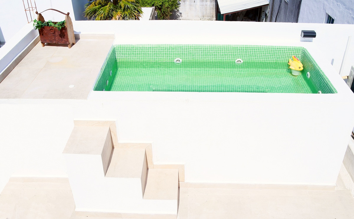 A private rooftop features a rectangular jacuzzi filled with green-tiled water, complemented by clean white walls. Steps lead up to the jacuzzi area, where a small rubber duck floats. A planter with greenery adds a touch of nature to the space.
