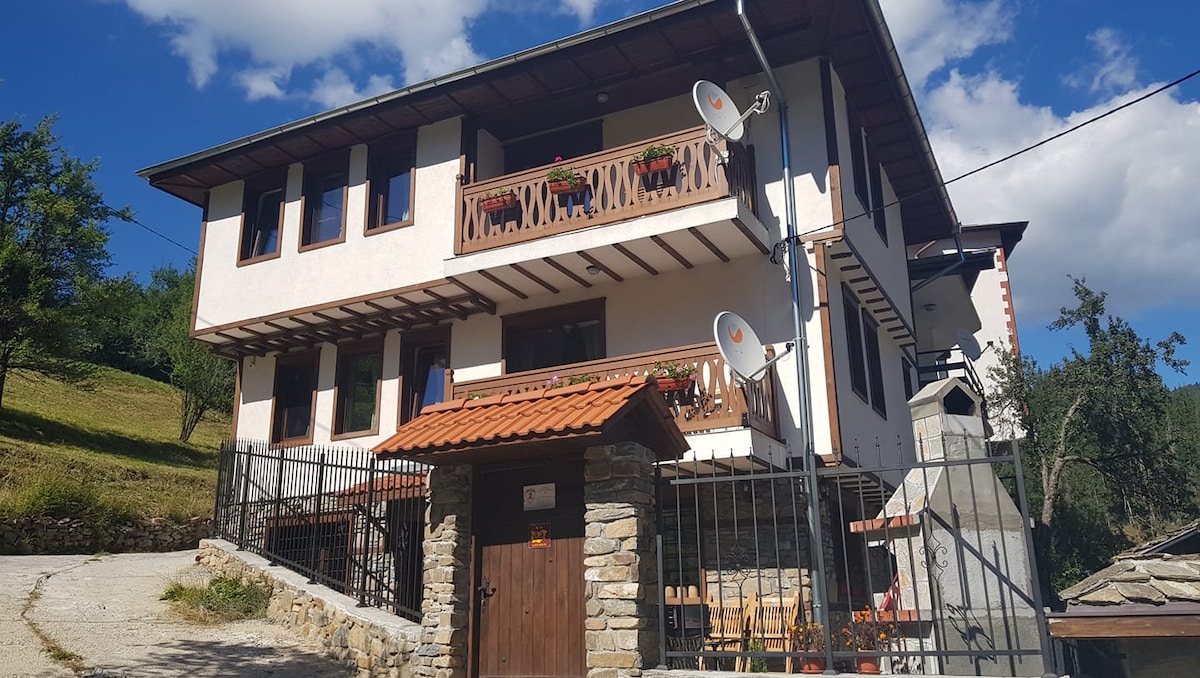 The exterior of a three-story house is depicted, showcasing a stone and wooden facade. Balconies adorned with flower boxes offer a welcoming touch. A gravel path leads to a wooden entrance, framed by a secure fence and surrounded by lush greenery and trees.