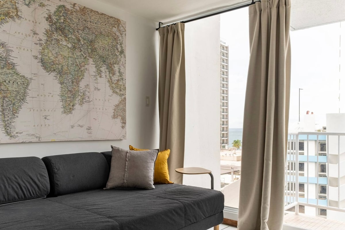 A comfortable seating area is showcased with a dark sofa adorned with decorative pillows. A world map hangs on the wall, providing a global touch. Natural light filters in through sliding doors that lead to a balcony, revealing views of the surrounding area.