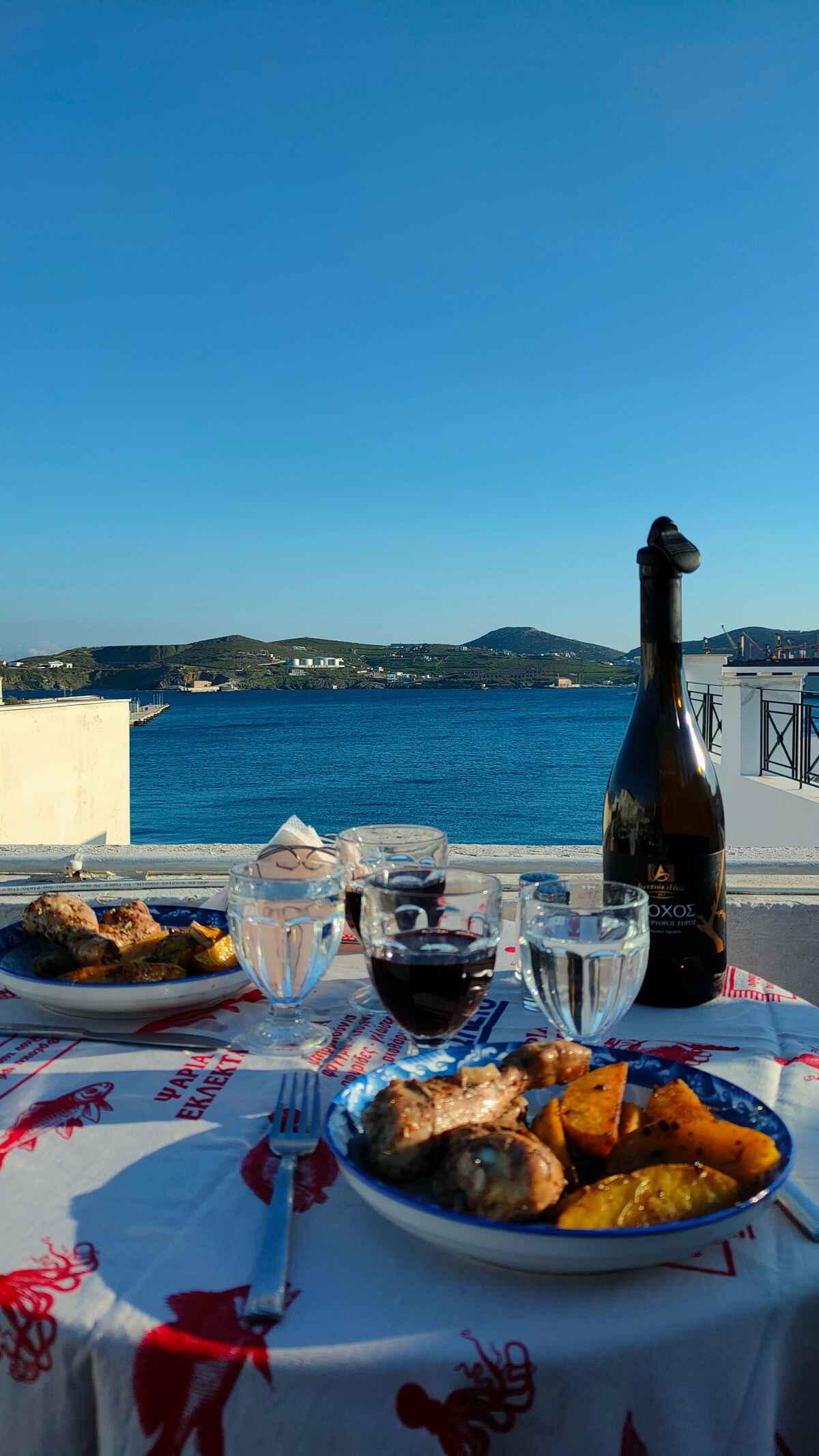 A dining table is set for two, featuring plates of grilled fish, roasted potatoes, and glasses of water and wine. A bottle of wine stands prominently. The backdrop showcases a panoramic view of the calm sea and distant hills under a clear blue sky.