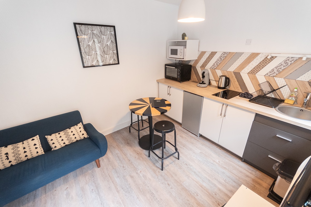A comfortable living area is depicted, featuring a blue sofa with patterned cushions. A round table with a unique design is set beside two black stools. The kitchenette displays modern appliances, including a microwave and kettle, against a stylish patterned backsplash.