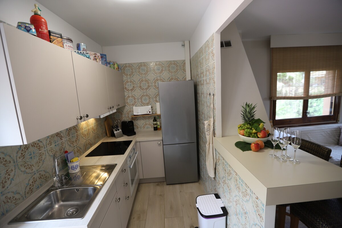 A modern kitchen features sleek cabinetry and a stainless steel refrigerator. Decorative tiles add character to the space, while natural light pours in through a window. A small dining area is visible, adorned with a fruit display and set with glassware.