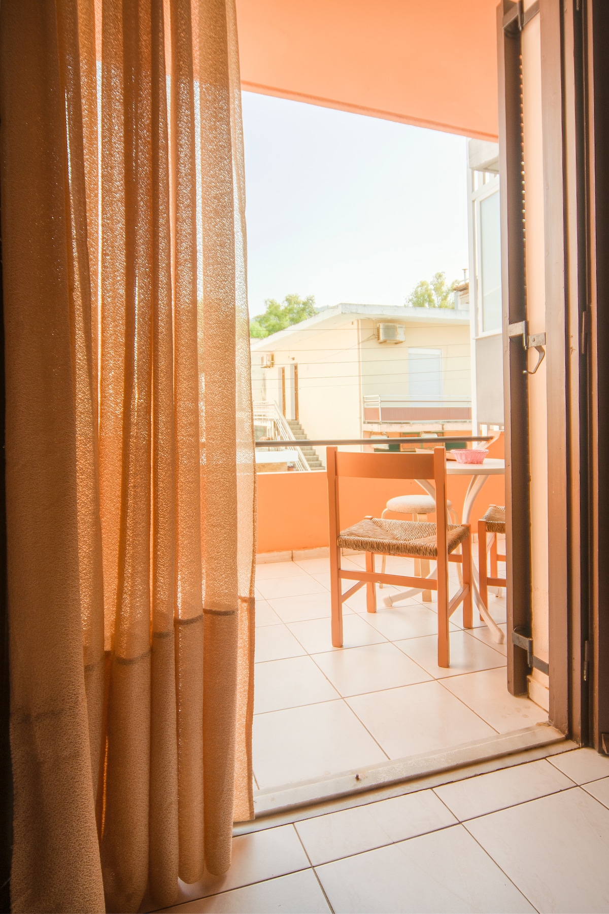 A view from inside the studio showcases a terrace area with a small table and two chairs. Light beige curtains frame the entrance, allowing natural light to enhance the warm tones of the space. The terrace overlooks the vibrant street beyond.
