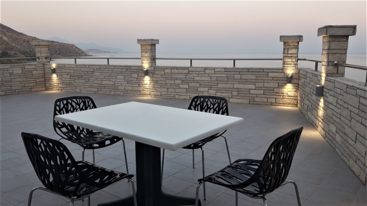 An outdoor terrace features a modern table surrounded by four contemporary chairs. Soft lighting illuminates the stone walls, creating a serene atmosphere with ocean views in the distance.