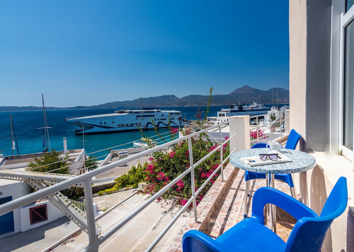A balcony area is equipped with a small round table and two blue chairs, offering a clear view of the port. Vibrant flowers adorn the railing, while boats are visible in the water under a clear blue sky.