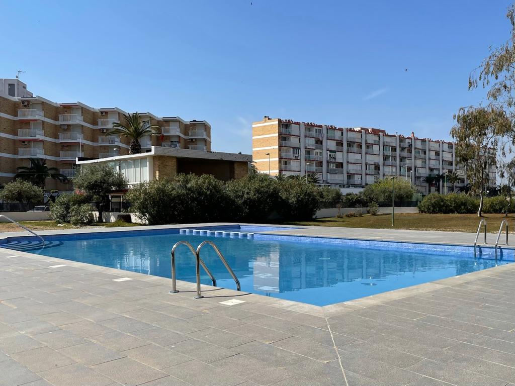 A clear swimming pool is visible, surrounded by well-maintained greenery and shrubs. Sun loungers are positioned nearby, with a spacious apartment building in the background. The scene is bright and captures the essence of outdoor relaxation on a sunny day.