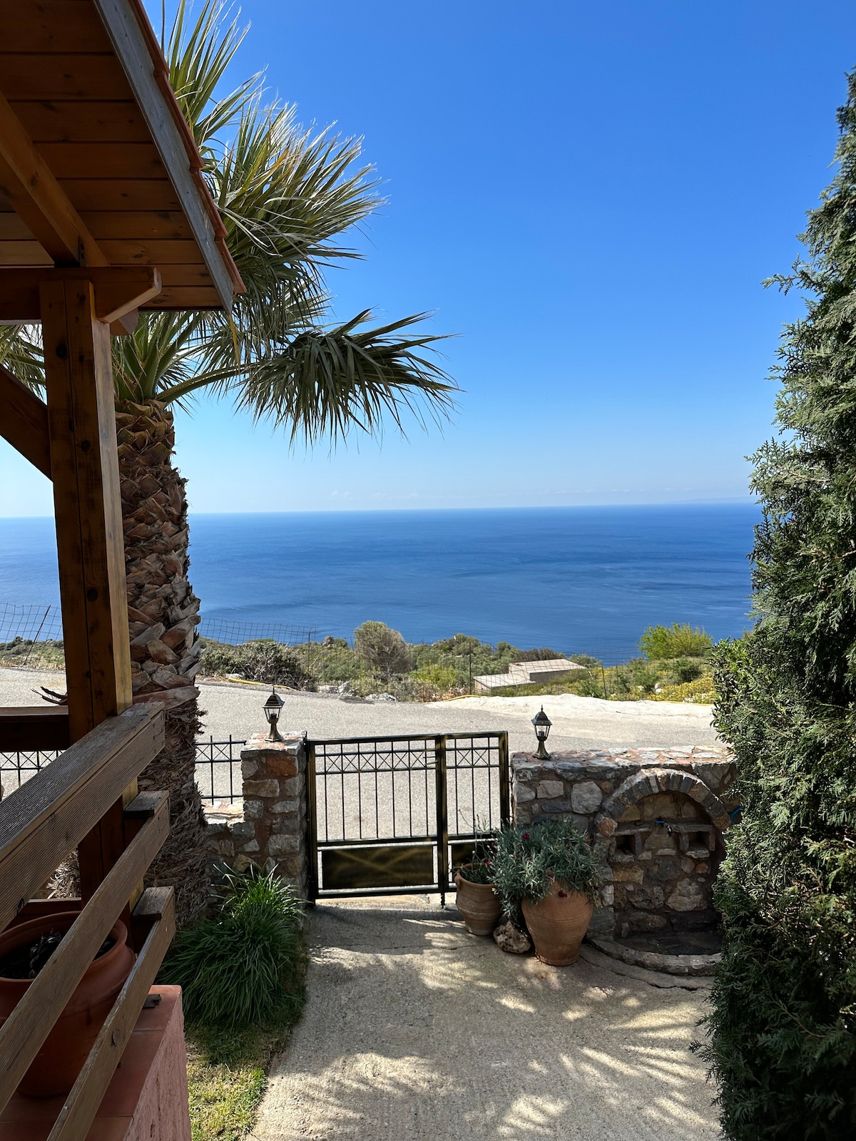 A welcoming entrance framed by palms and greenery opens to breathtaking views of the Libyan Sea. The path leads to a secure gate, surrounded by stone walls and decorative pots, enhancing the serene atmosphere of the villa's exterior.