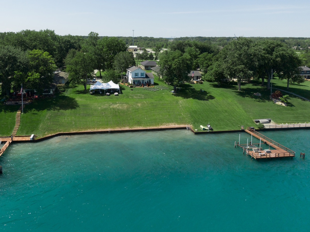 An aerial view captures the Hart's River House set on a green, spacious lawn by the water's edge. The vibrant turquoise water meets the shore, while a dock with small boats and seating areas is visible nearby. Surrounding trees offer shade and a natural backdrop.