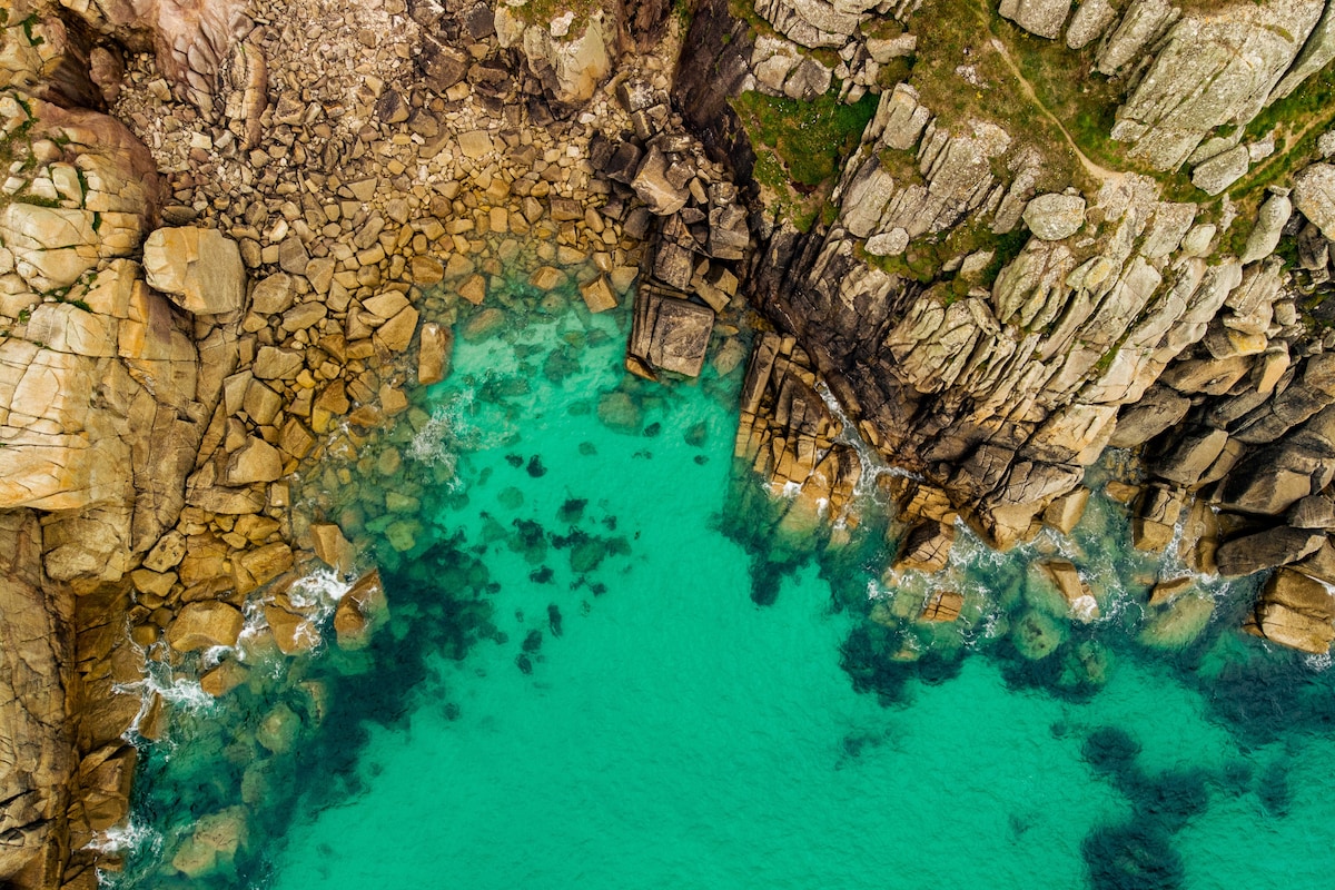 The image captures a striking coastal landscape featuring rocky cliffs and clear turquoise waters. A mix of large stones and smaller pebbles is visible along the shore, while the ocean reveals rocky underwater terrain, creating a vibrant contrast against the calming water.