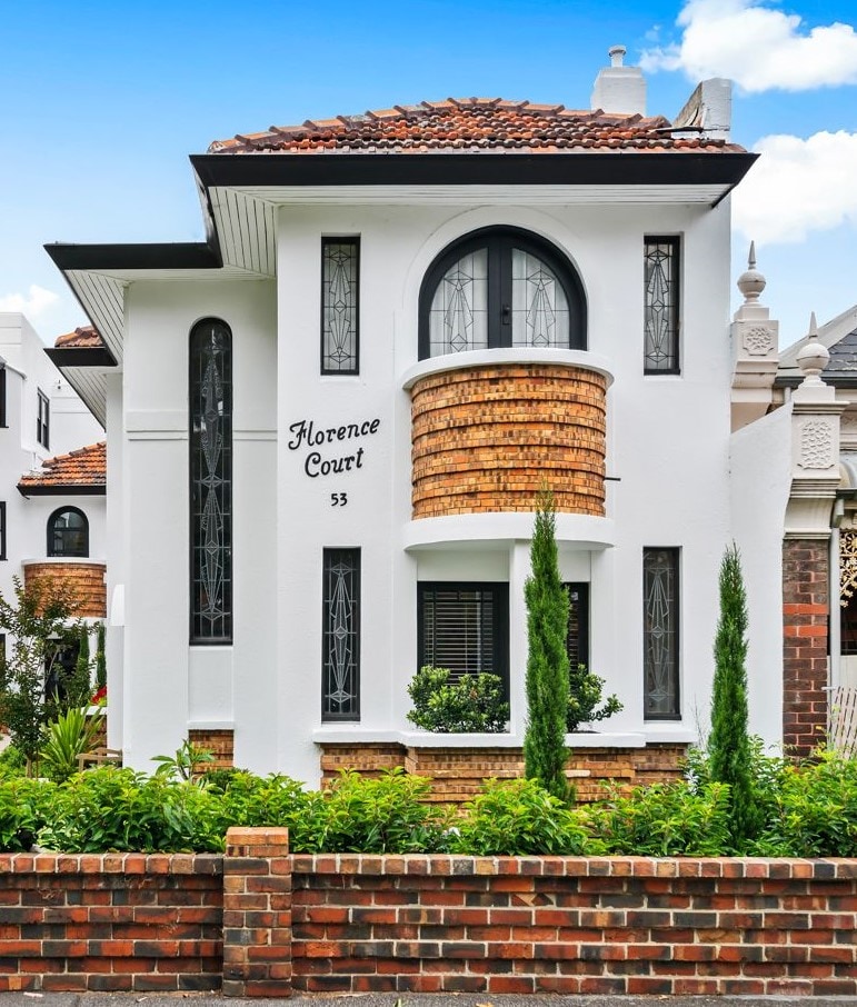 The exterior of Florence Court displays distinctive Art Deco architecture with elegant curved lines and a textured facade. Tall windows with lead light accents complement the brick detailing, while neatly landscaped greenery surrounds the entrance, enhancing the inviting appearance of the building.
