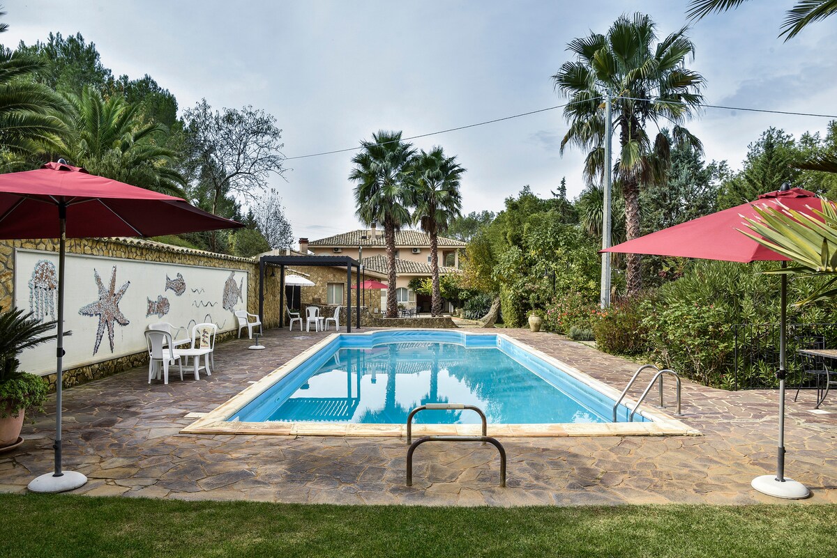 A serene outdoor area features a swimming pool surrounded by palm trees and greenery. Two red umbrellas shade white lounge chairs adjacent to the pool, while a stone pathway provides access. A decorative wall with sea-themed motifs enhances the relaxing atmosphere.