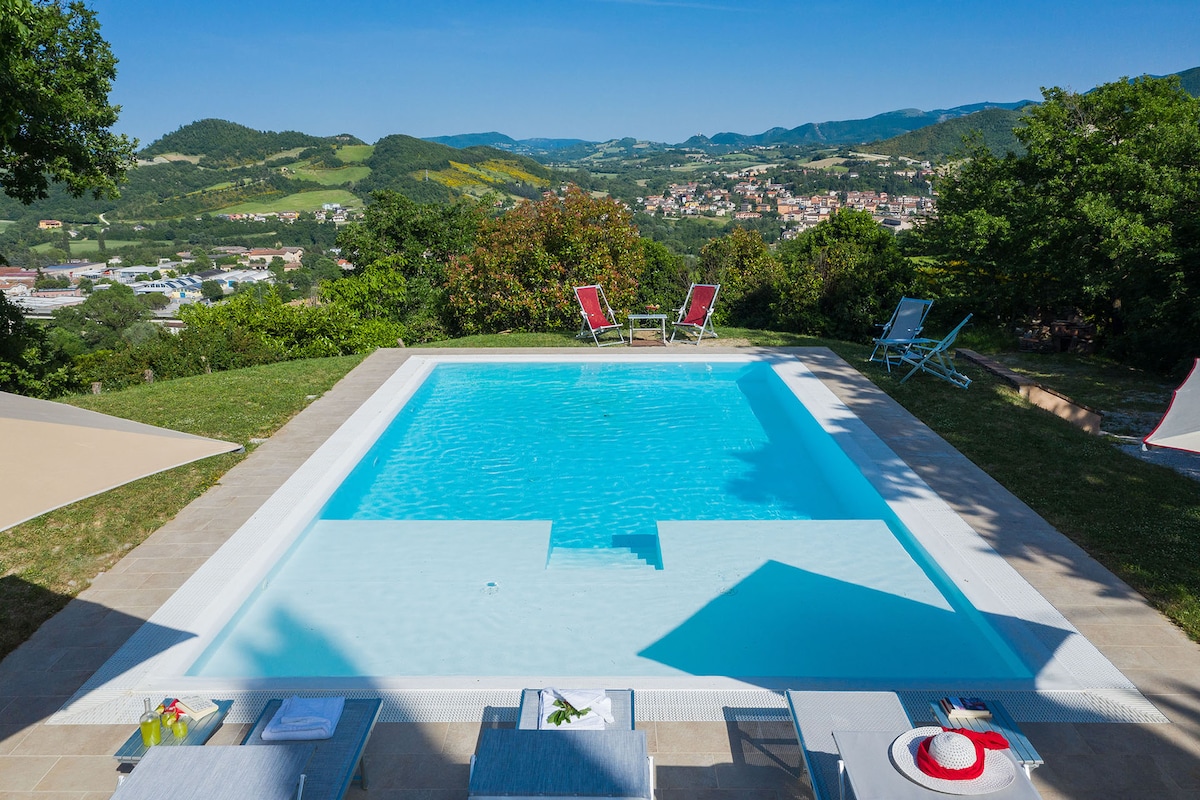 A clear swimming pool, 10 x 5.5 meters, sits within a landscaped area, surrounded by lounge chairs. The view encompasses rolling hills and a distant town under a clear blue sky, creating a serene atmosphere for relaxation.