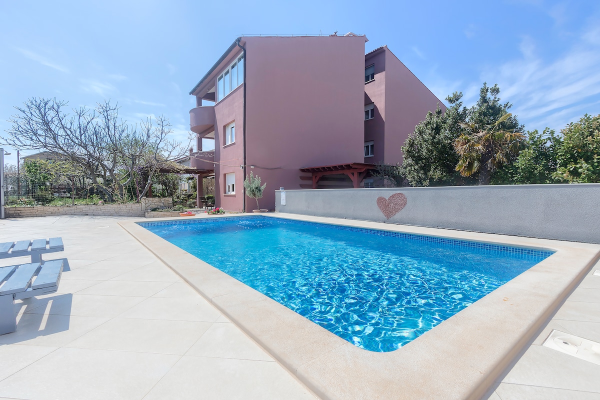 A private swimming pool is surrounded by a tiled patio, providing ample space for relaxation. Lush greenery is visible in the background, and a light-filled building with multiple levels stands nearby. A shaded seating area is included for guest comfort.