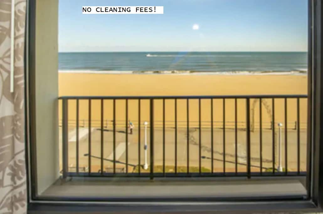A view from a balcony showcases the sandy beach and ocean beyond. The horizon meets the water under a clear blue sky, with footsteps visible on the adjacent walkway. A railing provides a seamless transition between indoor and outdoor landscapes.