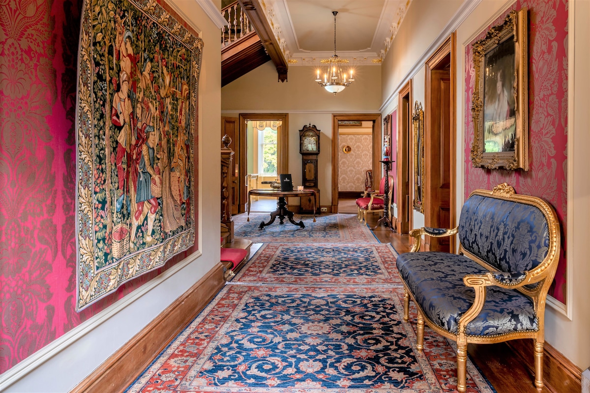 A grand hallway features intricate patterned wallpaper and elegant woodwork. A vintage-style blue settee with gold accents invites seating. Richly colored rugs and a tapestry add visual warmth, while soft light filters through windows, illuminating the space leading to various rooms.