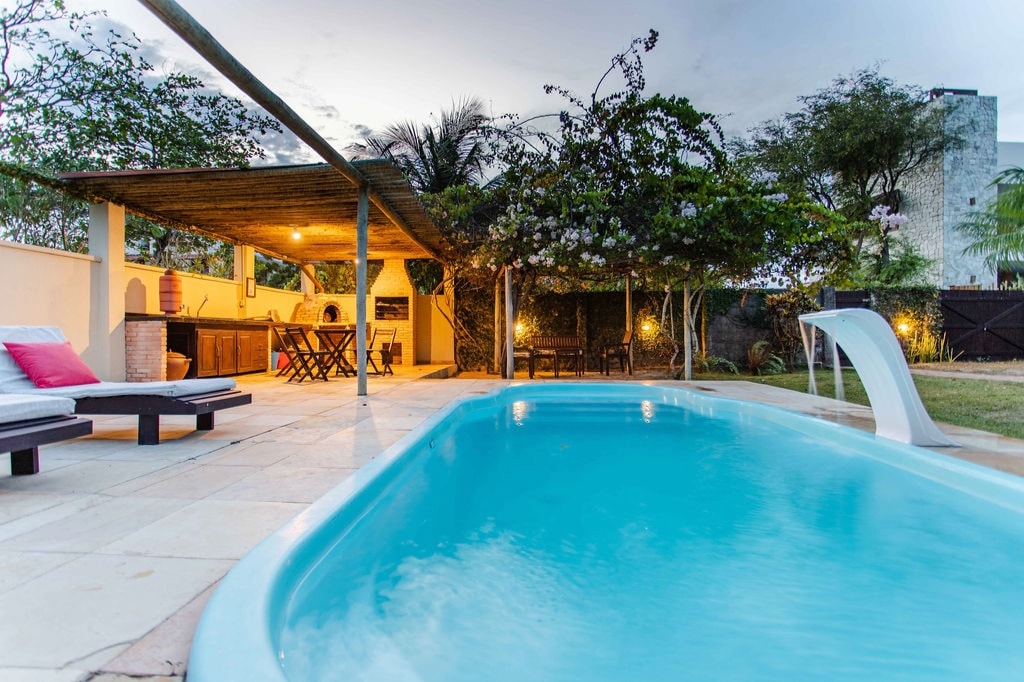 A private swimming pool is surrounded by a stone deck and equipped with a comfortable lounge area featuring striped seating. An outdoor kitchen and dining space are illuminated softly, nestled beneath wooden beams and surrounded by lush greenery.