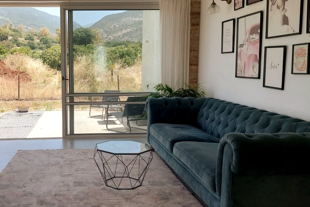 A spacious living area features a stylish tufted sofa in muted teal, complemented by a geometric coffee table. Large glass doors open to picturesque views of the surrounding hills, while light filters in through sheer curtains, enhancing the bright and airy ambiance.
