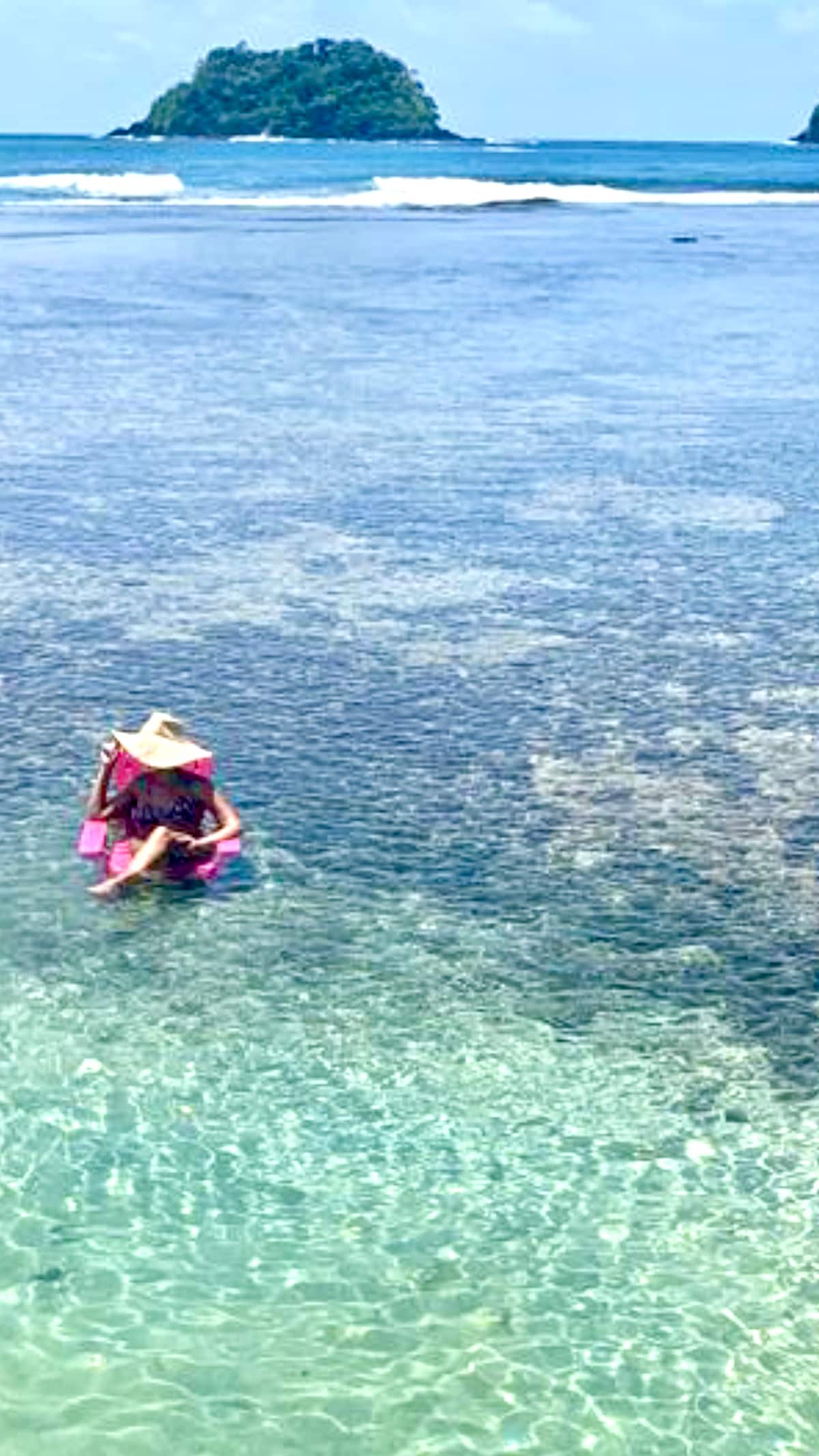 Clear turquoise waters are shown, with gentle waves and a sandy bottom visible through the water. A person floats on a pink raft, wearing a wide-brimmed hat. In the distance, a lush green island contrasts against the blue sky.