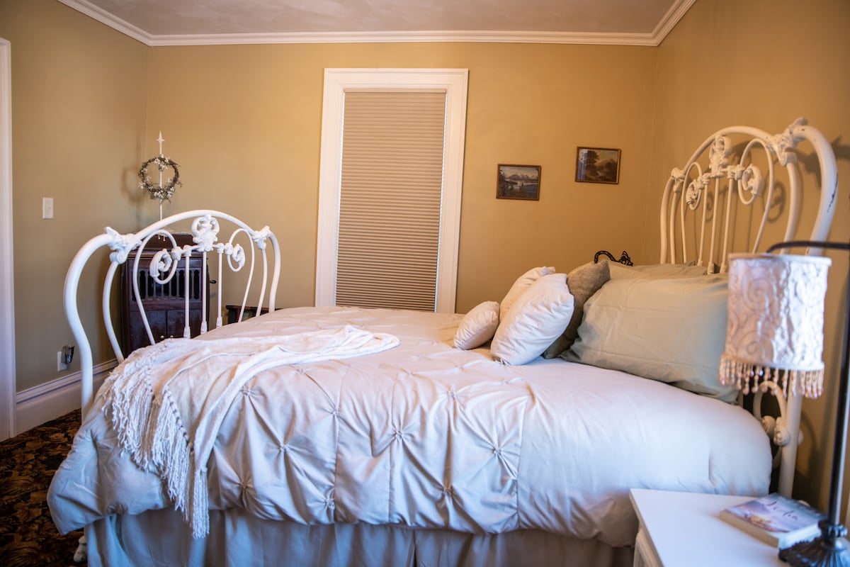 A charming bedroom showcases a white iron bed adorned with plush linens and an assortment of pillows. Soft hues grace the walls, while a decorative lamp provides gentle light. A small bedside table complements the space, enhancing the cozy atmosphere of the room.