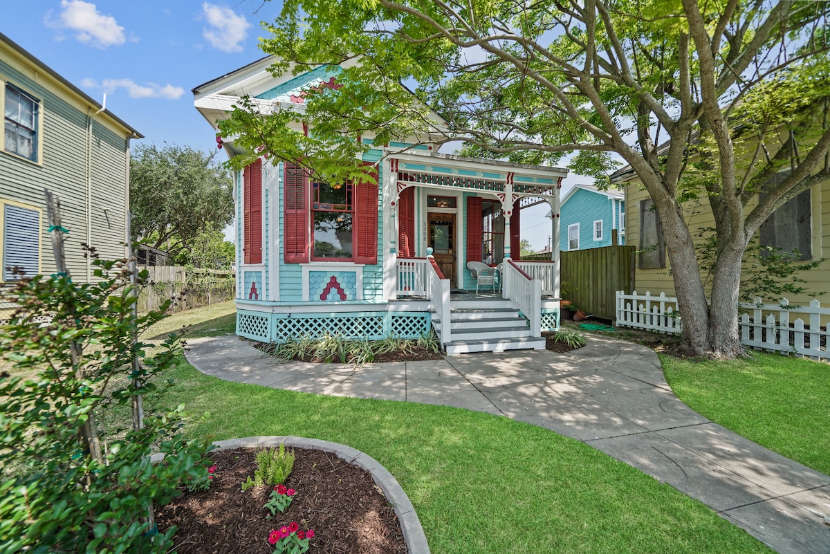 The exterior of a charming Victorian cottage is showcased, featuring intricate woodwork and vibrant colors. A pathway leads to the inviting front porch, surrounded by well-maintained greenery and flower beds. Large windows allow ample natural light, and a shady tree provides a relaxing outdoor space.
