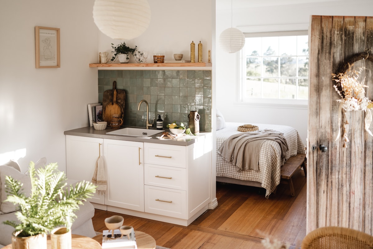 A compact kitchen is visible, featuring white cabinetry and a gray countertop. Decorative items are arranged on a wooden shelf above the sink. The adjoining sleeping area has a cozy bed with light bedding, complemented by a window offering a view of the outdoors.