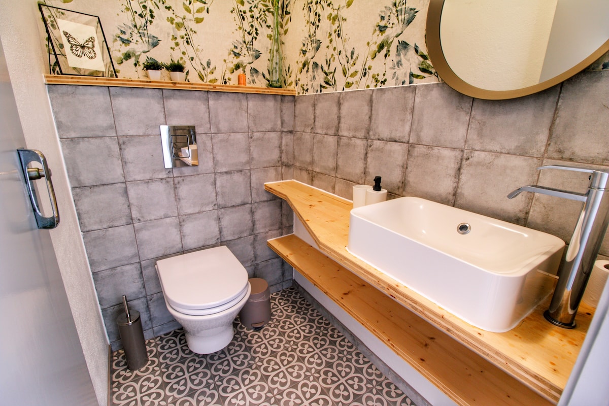 A modern bathroom features a wall-mounted toilet and a stylish sink with a rectangular basin. Wooden shelves provide storage space, and a round mirror reflects natural light. Floral wallpaper enhances the space, while patterned tiles provide a decorative flooring element.