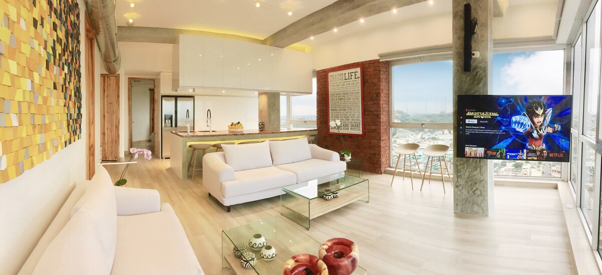 A spacious living area features a comfortable white sofa and a glass coffee table. Natural light floods in through large windows, providing stunning views of the city and river. A flat-screen TV is mounted on the wall, and the modern kitchen is visible in the background.