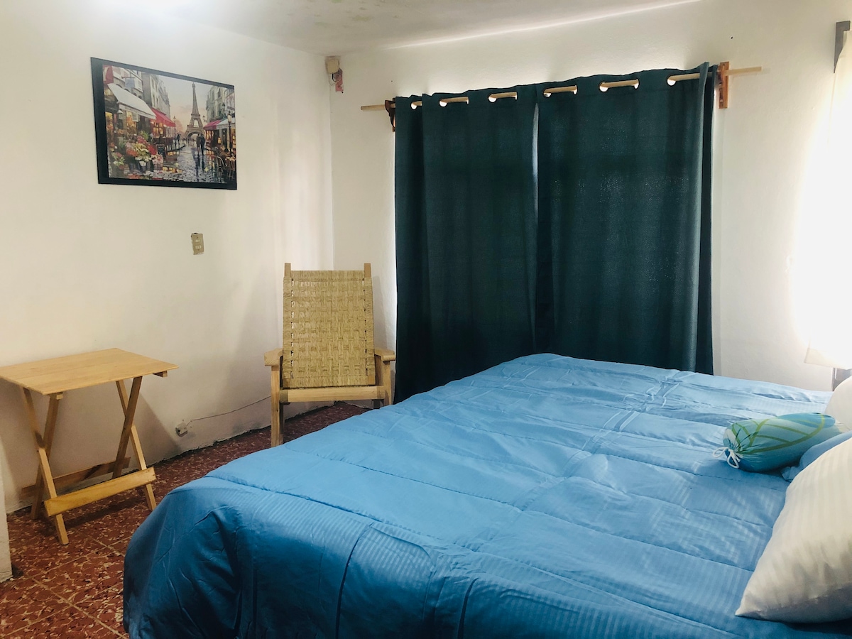 A spacious sleeping area features a large bed with a blue bedspread and a decorative green towel. A wooden chair and a simple table are visible nearby. Dark curtains frame the window, allowing natural light to softly illuminate the room. A wall-mounted artwork adds a touch of color.