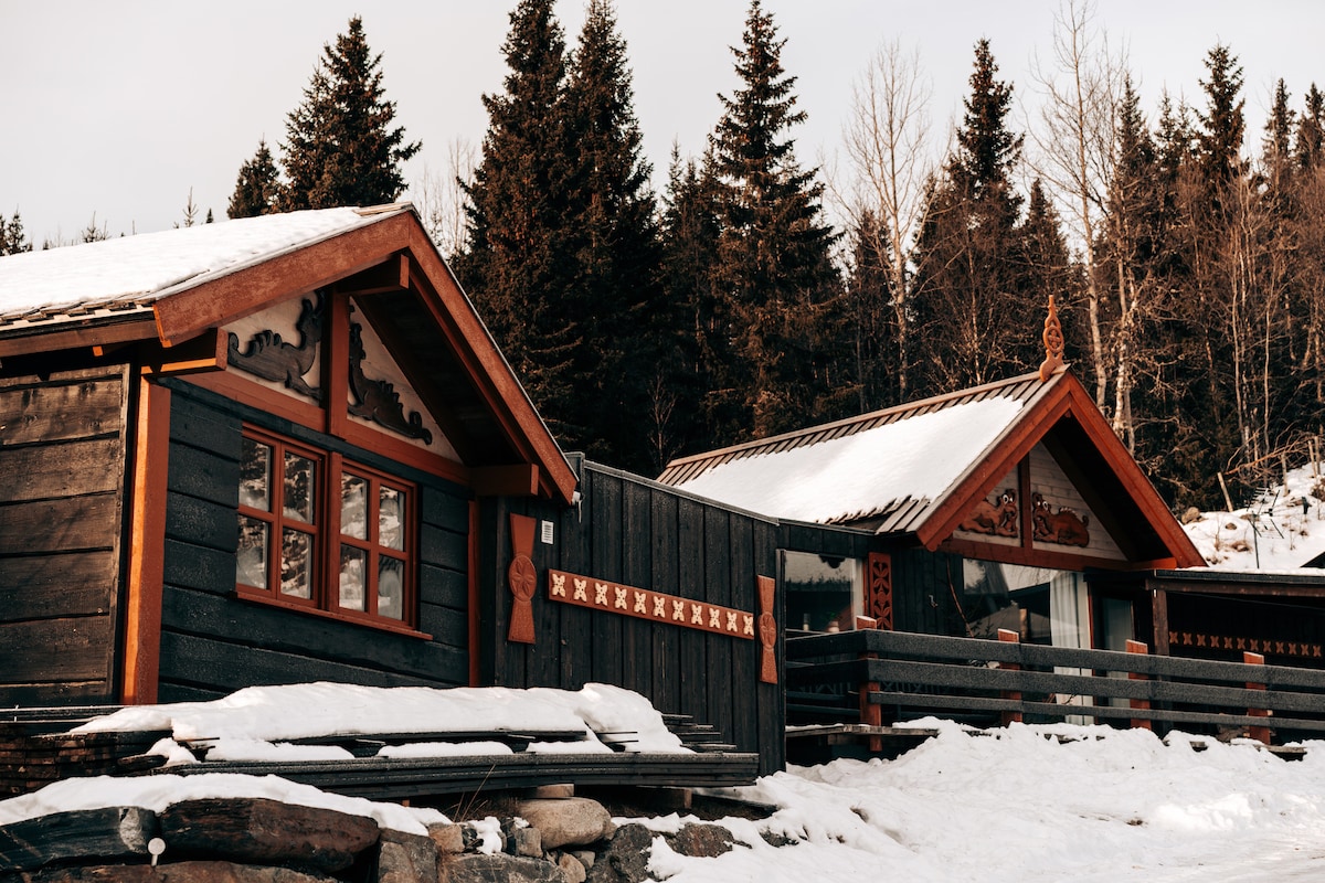 Two traditional-style cottages are set against a backdrop of evergreen trees and snow-covered ground. The wooden exteriors feature intricate carvings and decorative trims. Sunlight casts gentle shadows on the snow, enhancing the rustic charm of the structures.