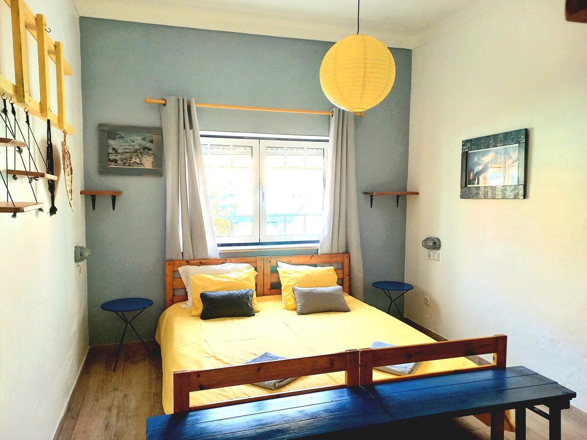 A bright double room features a spacious bed dressed in yellow linens, flanked by two small blue tables. Natural light fills the room through a window, adorned with white curtains, and a paper lantern hangs from the ceiling. Wooden shelves are mounted on the walls, adding to the decor.