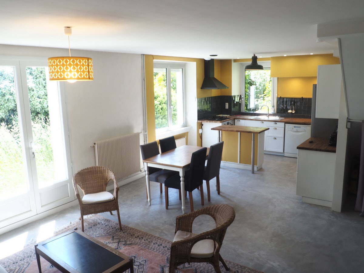 A spacious living area features a dining table surrounded by black chairs and a cozy seating arrangement with woven chairs. A kitchen is visible in the background, showcasing modern appliances and a warm yellow accent wall. Light floods in through large windows offering views of the garden.