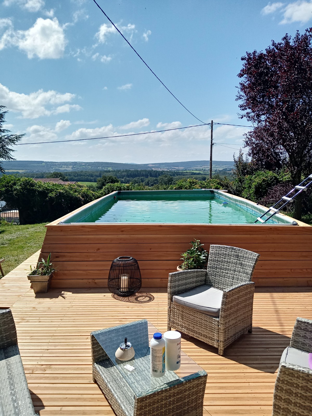 A wooden deck features wicker chairs arranged around a small table, offering a comfortable outdoor seating area. In the background, a rectangular pool reflects the sky, surrounded by greenery and distant hills. A single lantern and potted plants add subtle decor.