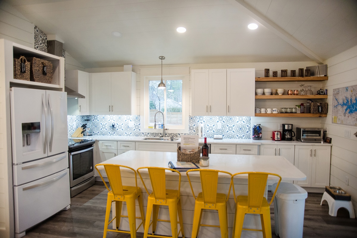 A modern kitchen features a spacious island with yellow stools and white cabinetry. Decorative tiles are highlighted on the backsplash, while shelves display an assortment of kitchenware. Natural light illuminates the area through the window above the sink, enhancing the inviting workspace.
