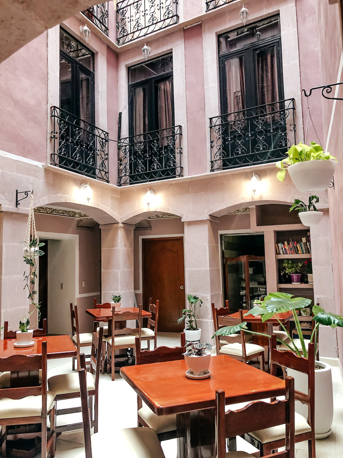 An inviting dining area features several wooden tables surrounded by light-colored chairs. Ornamental plants are placed throughout the space, and a cozy nook with shelves is visible in the background. Large windows with dark drapes allow natural light to enhance the ambiance.
