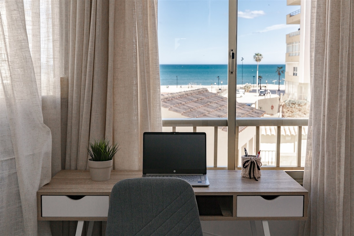 A workspace is displayed with a laptop on a light-colored desk, positioned near a large window that offers a view of the sea. Light drapes frame the window, allowing natural light to enter the room, while a small plant adds a touch of greenery.