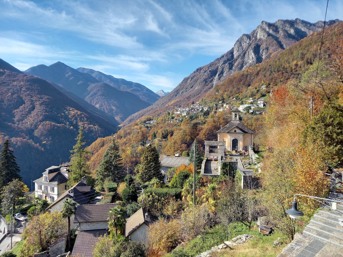 A scenic view showcases the surrounding alpine landscape, featuring rolling hills and vibrant autumn foliage. The mountains rise prominently in the background, while charming homes with outdoor terraces are positioned throughout the valley, under a clear blue sky.