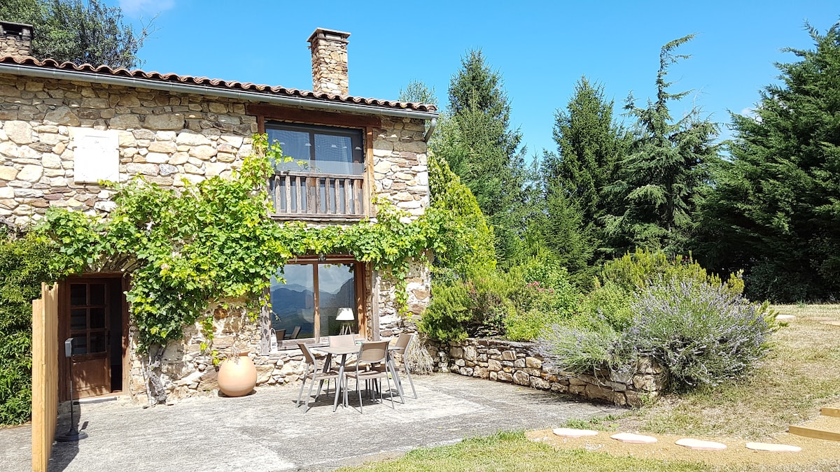 A rustic stone building is surrounded by trees and greenery, featuring a balcony with wooden railings. An outdoor dining area with a table and chairs is set on a patio adorned with decorative plants, offering a view of the natural landscape.