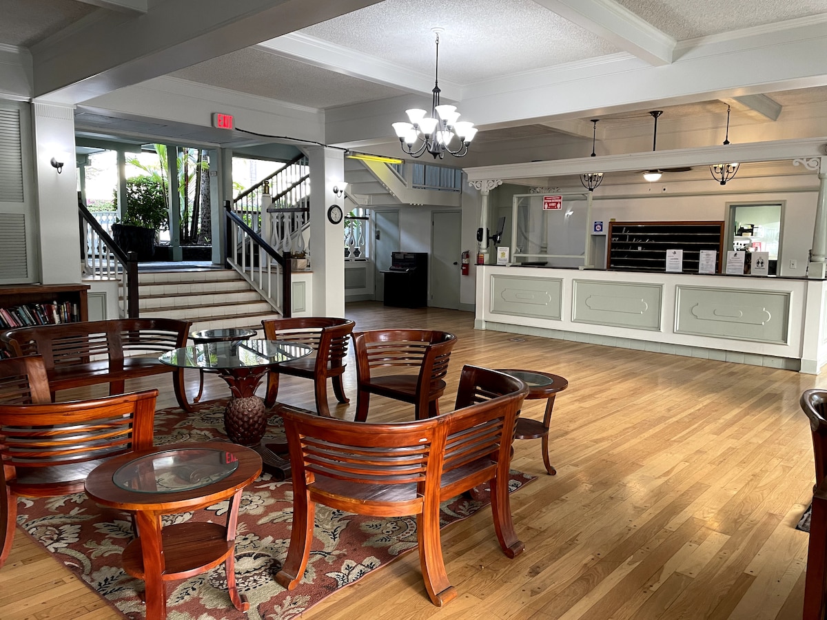 A spacious lobby area features polished wooden floors and a central seating arrangement consisting of multiple wooden chairs and tables. Natural light filters in through large windows, illuminating the reception desk, which is situated against the wall. Potted plants add greenery to the inviting space.