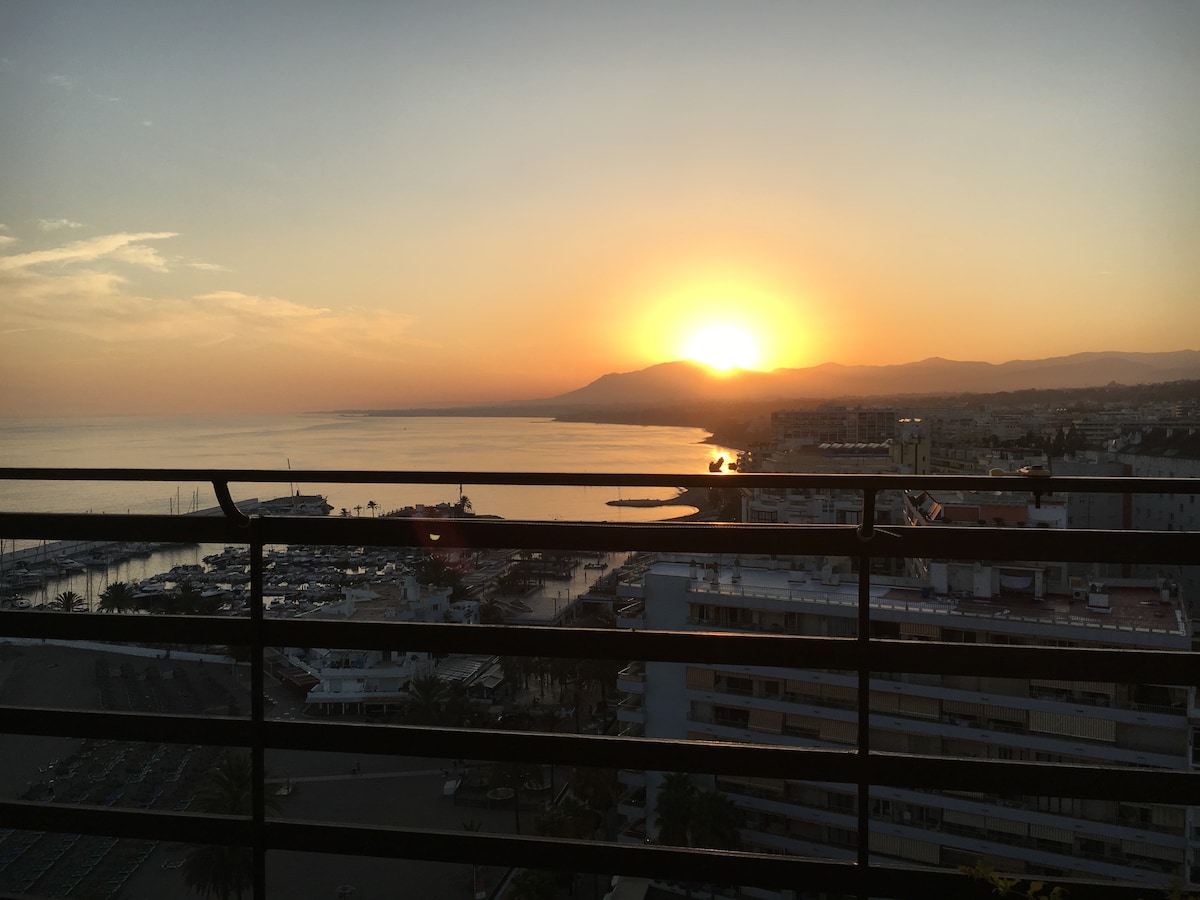 A distant view of a sunset is captured over the horizon, with warm orange and yellow hues blending into the sky. The coastline and mountains are visible, alongside the marina and city below from a terrace railing.