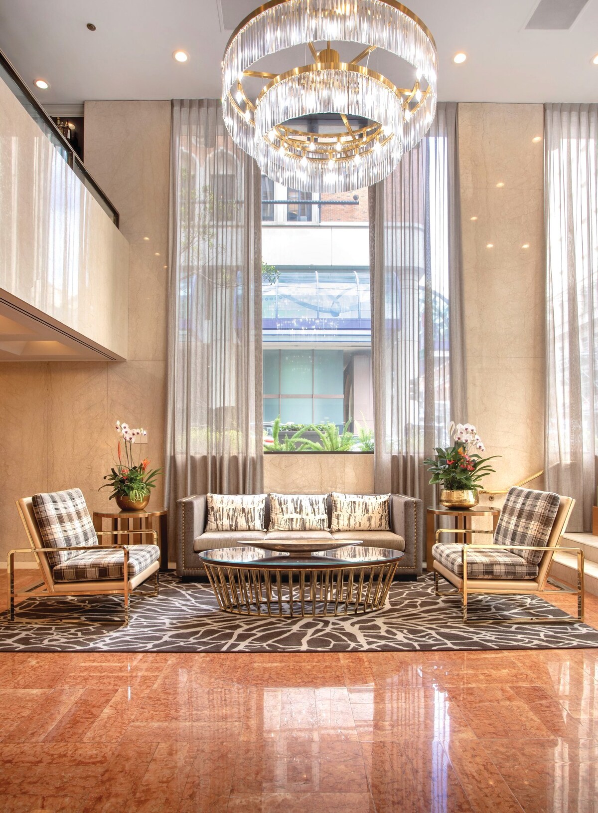 A stylish lobby area features three plaid armchairs and a circular coffee table set on an elegant rug. Large windows allow natural light to filter through sheer curtains, while a modern chandelier hangs above, providing a warm ambiance. Potted plants add a touch of greenery.