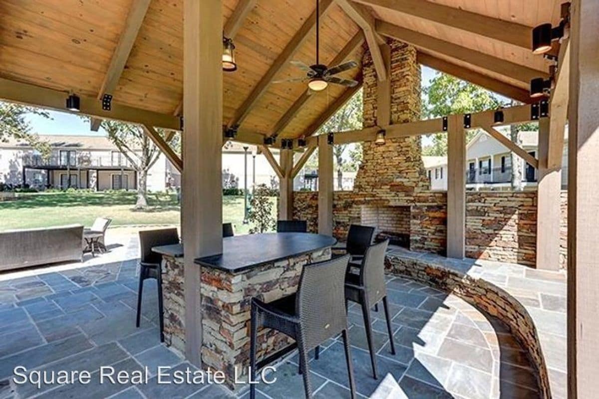An outdoor pavilion features a stone wall with a built-in counter area. Several dark wicker chairs are arranged around the counter, under a wood-beamed ceiling adorned with ceiling fans. The surrounding green lawn is visible in the background.
