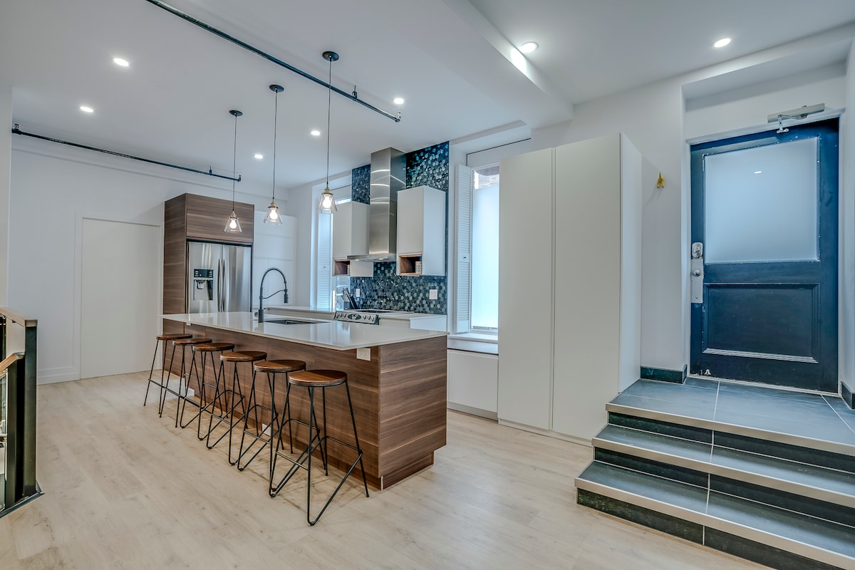 The kitchen area features a large island with several high-backed stools, providing ample space for gathering. Modern appliances are integrated into sleek cabinetry, while natural light enhances the welcoming atmosphere. The entryway shows a stylish door, complemented by a staircase leading to the upper level.